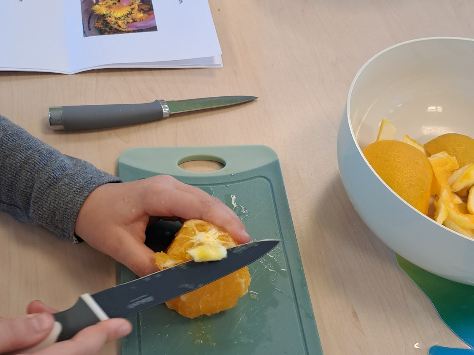 Person schneidet eine Orange auf einem Schneidebrett, daneben liegt ein offenes Rezeptbuch.