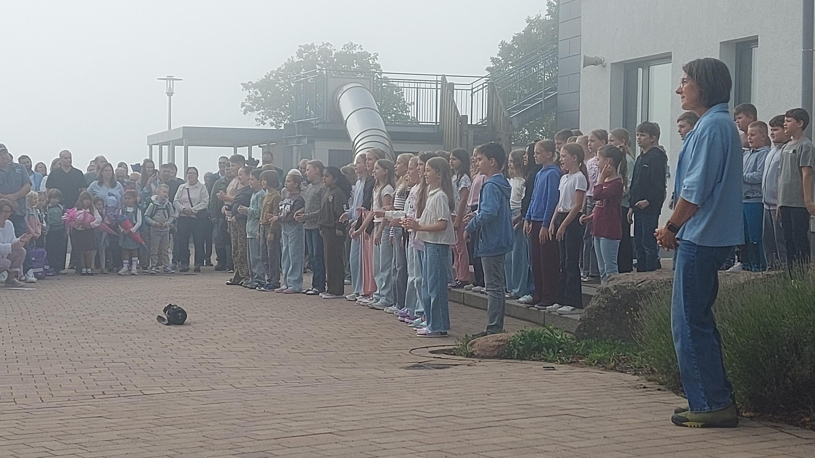 Kindergruppe, die vor einem Publikum im Freien auftritt, mit einem Erwachsenen daneben.