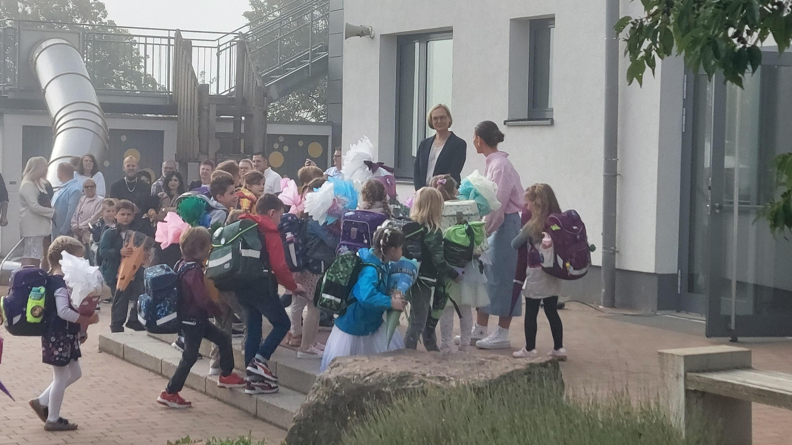 Kinder mit Rucksäcken und Schultüten vor einer Schule, umgeben von Erwachsenen.