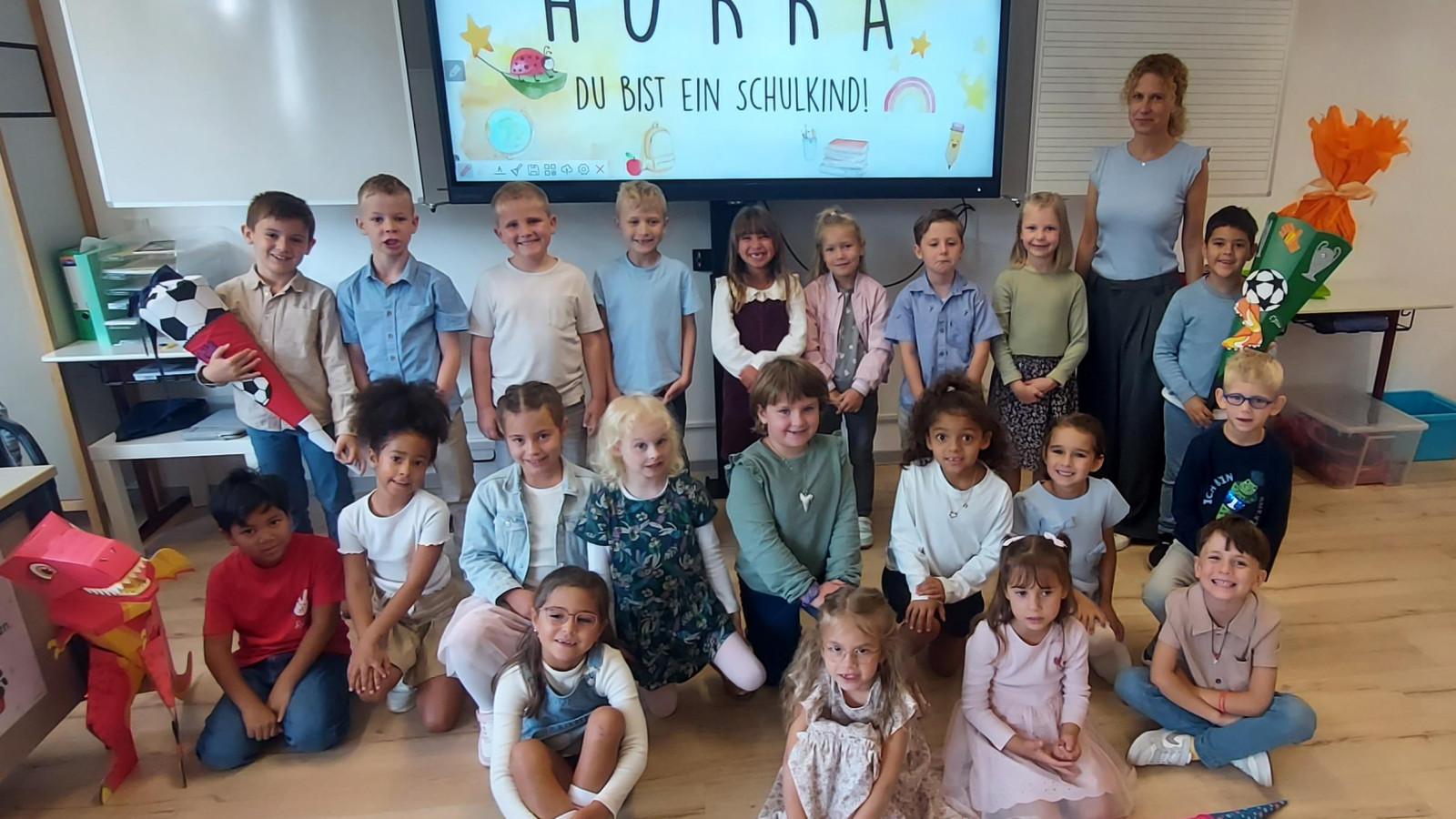 Gruppe von Kindern und Lehrerin vor einer Tafel mit der Aufschrift 'HURRA DU BIST EIN SCHULKIND!'