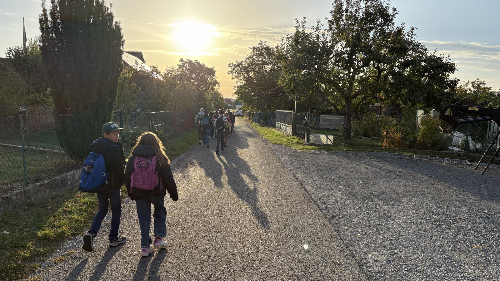 Zwei Kinder mit Rucksäcken spazieren auf einer sonnigen Straße in Richtung Sonnenuntergang.