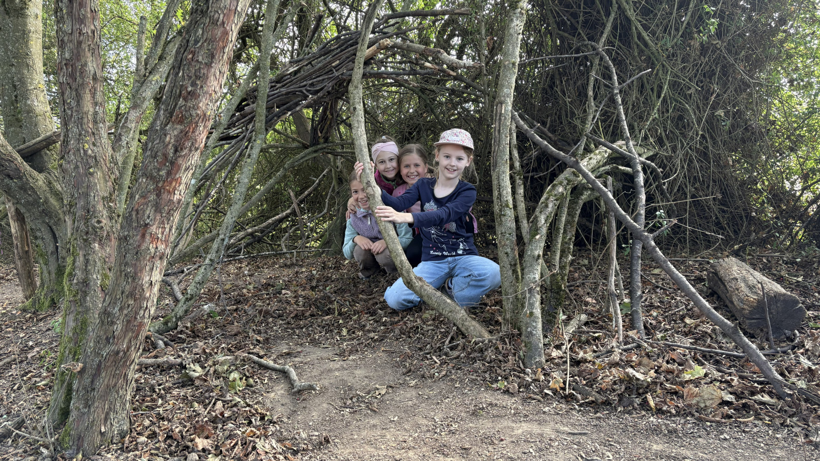 Drei lachende Kinder in einer aus Ästen gebauten Hütte im Wald.