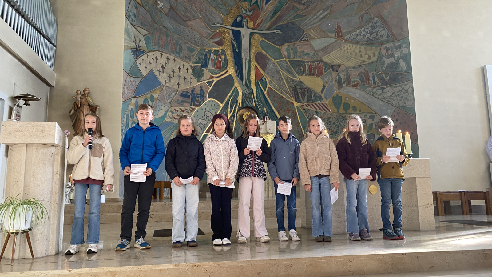 Kinder stehen in einer Reihe vor einem Wandgemälde in einer Kirche und halten Zettel.