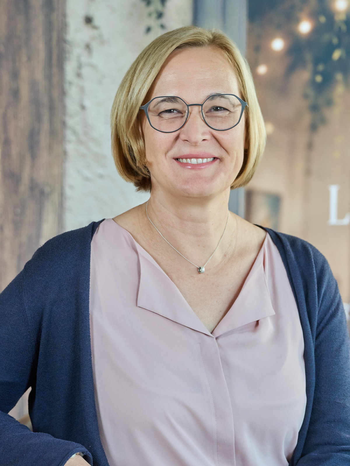 Eine lächelnde Frau mit Brille in rosa Bluse und dunklem Cardigan.