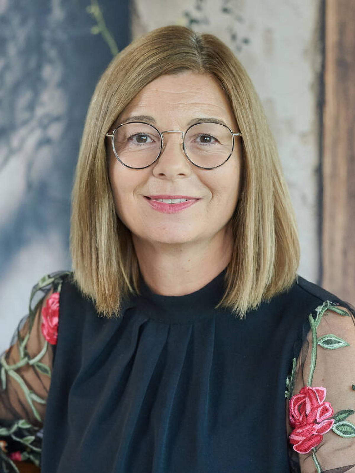 Frau mit Brille in schwarzem Kleid mit floralen Ärmeln vor rustikalem Hintergrund.
