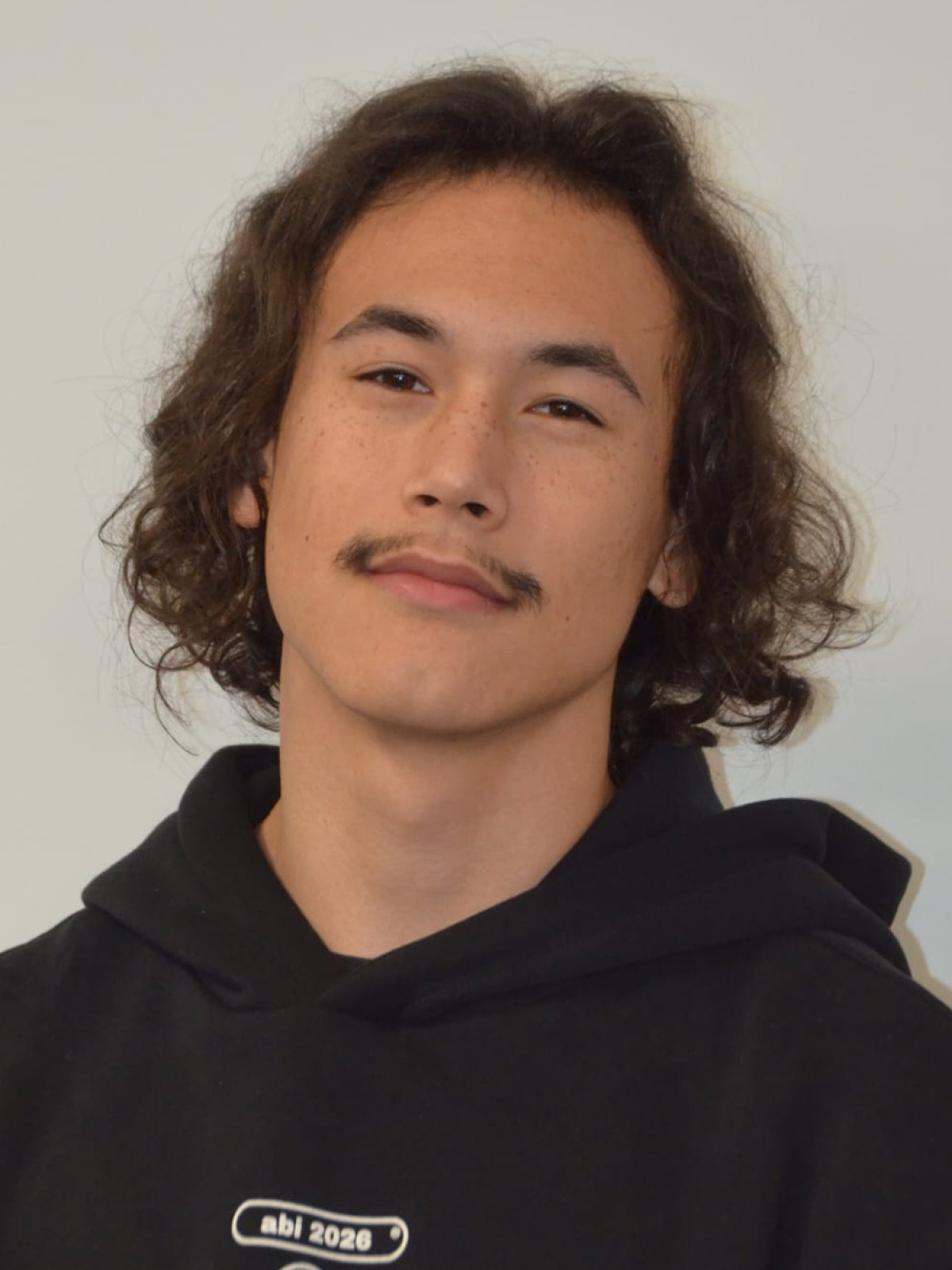 Junger Mann mit lockigem Haar und Schnurrbart, trägt schwarzen Hoodie, neutraler Hintergrund.