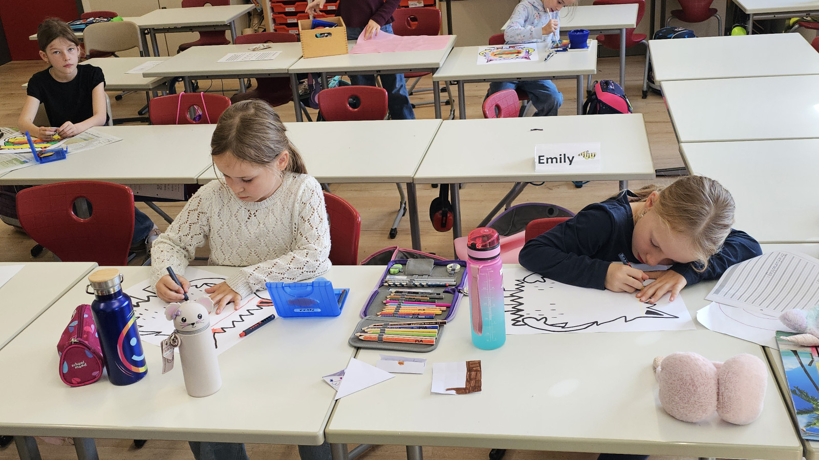 Kinder sitzen an Tischen im Klassenzimmer und arbeiten an Zeichnungen, umgeben von Malutensilien.