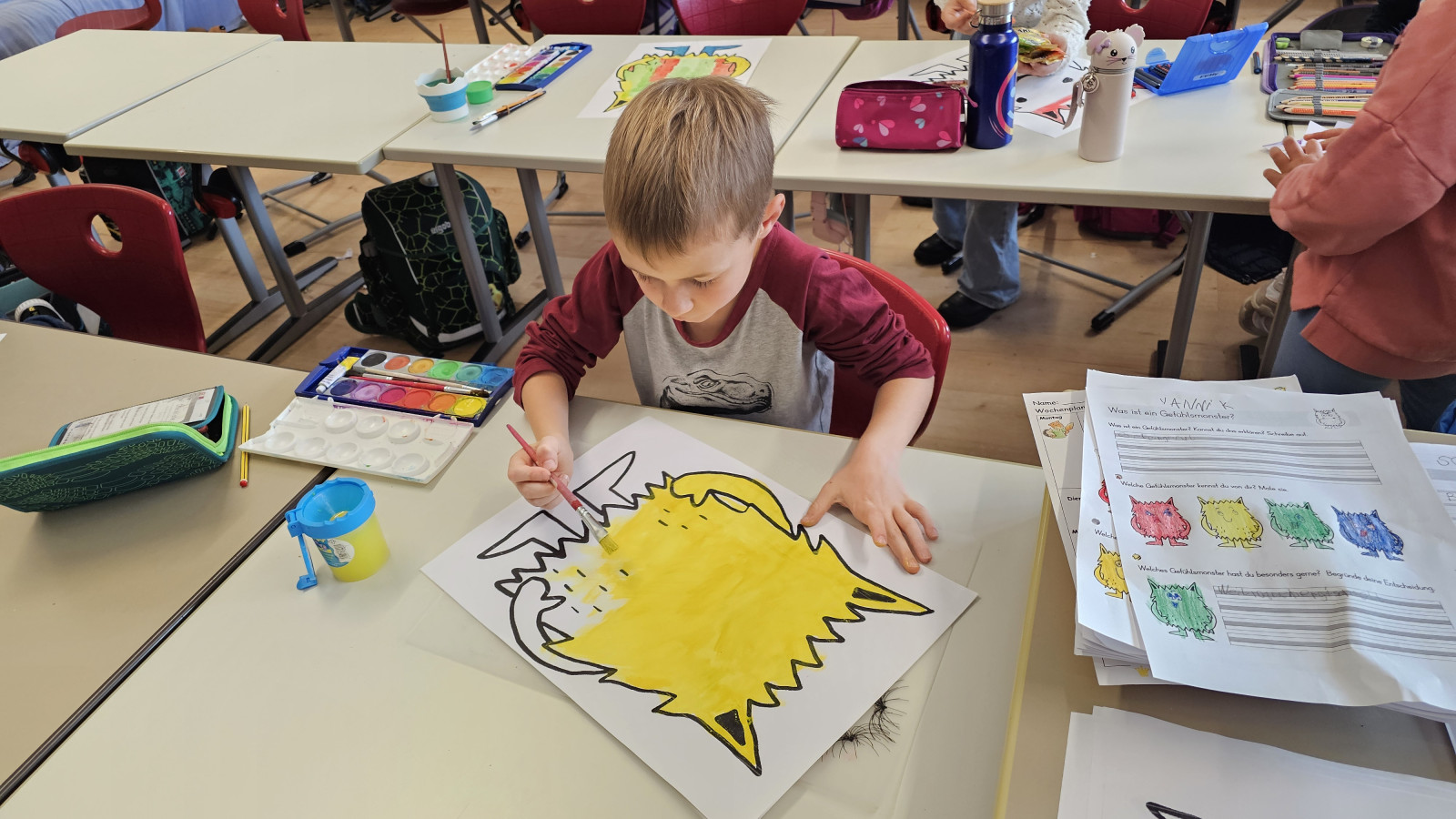 Junge im Klassenzimmer malt mit Wasserfarben ein Bild aus.