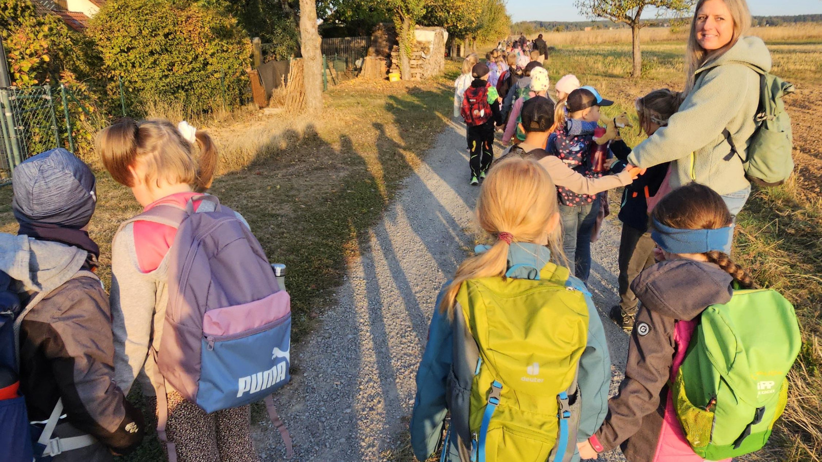 Kinder mit Rucksäcken gehen auf einem Feldweg, während die Sonne untergeht.