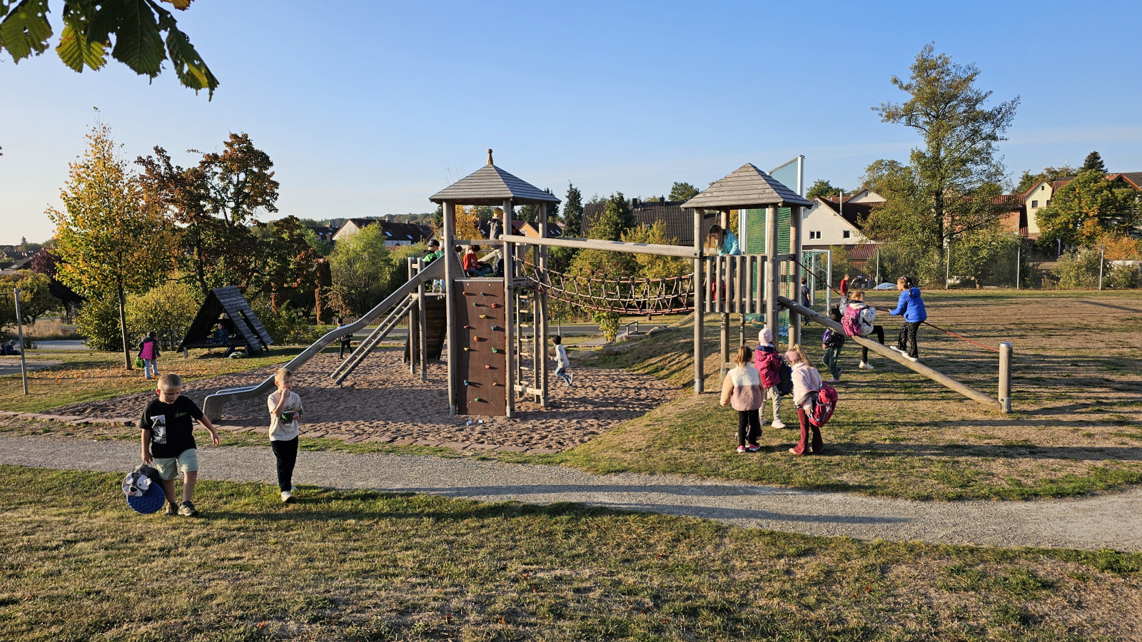 Kinder spielen auf einem gut besuchten Spielplatz an einem sonnigen Tag, umgeben von Grünflächen.