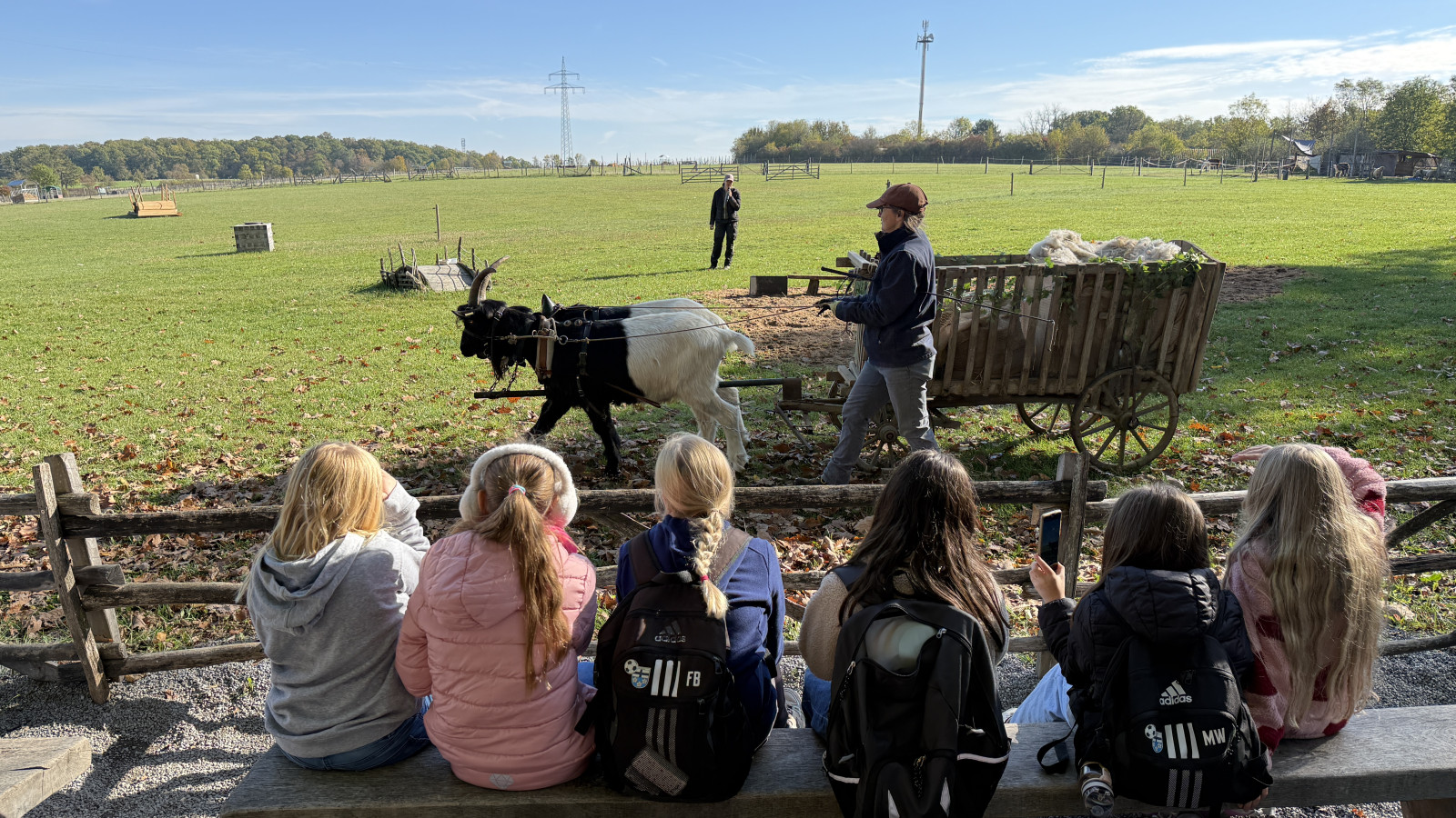 Kinder sitzen auf einer Bank vor einem Feld, wo eine Ziege einen Karren zieht, geführt von einer Person.
